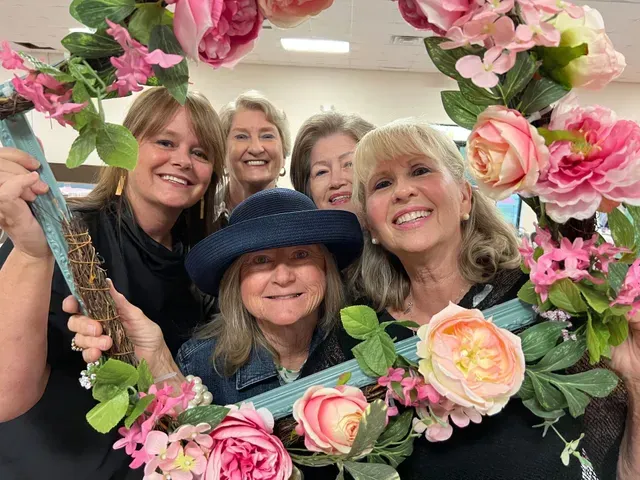 Five smiling people posing together behind a large, decorative wreath adorned with pink and peach faux flowers.