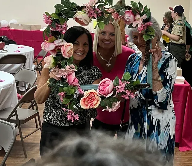 Three smiling people pose together holding a decorative wooden frame adorned with artificial pink flowers in a banquet hall.