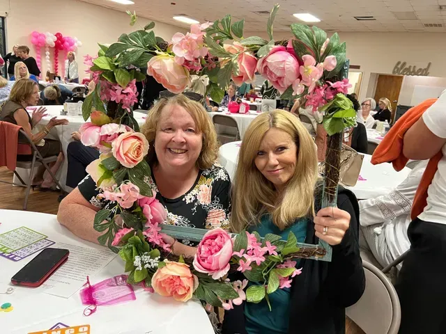Two people smiling behind a large, rectangular flower-covered frame in a room decorated for a social event.