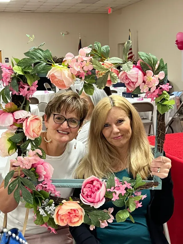 Two smiling women hold a large, rectangular wooden frame decorated with colorful artificial flowers.
