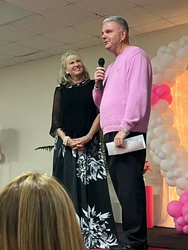 A person in a pink sweater speaks into a microphone while a person in a black patterned dress smiles beside them.