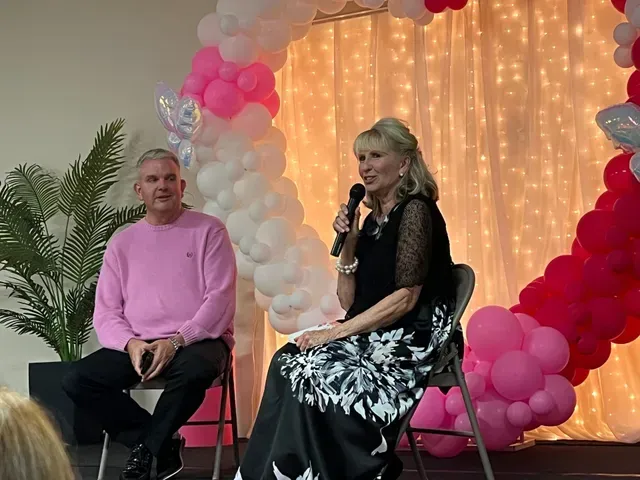 Two people sit on chairs on a stage in front of a backdrop of pink and white balloons and a curtain with fairy lights.