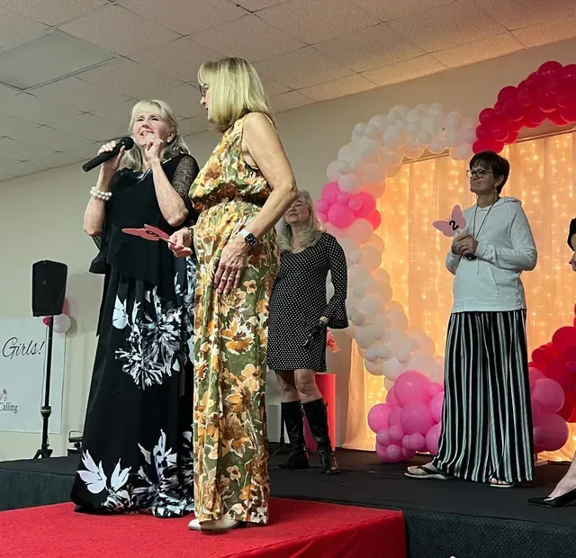 Four people stand on a stage with a red carpet and a large heart-shaped balloon arch behind them. One is speaking.