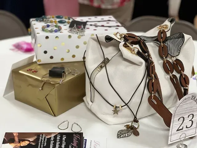 A white handbag with a brown-linked strap sits on a table next to wrapped gift boxes and jewelry on a white tablecloth.