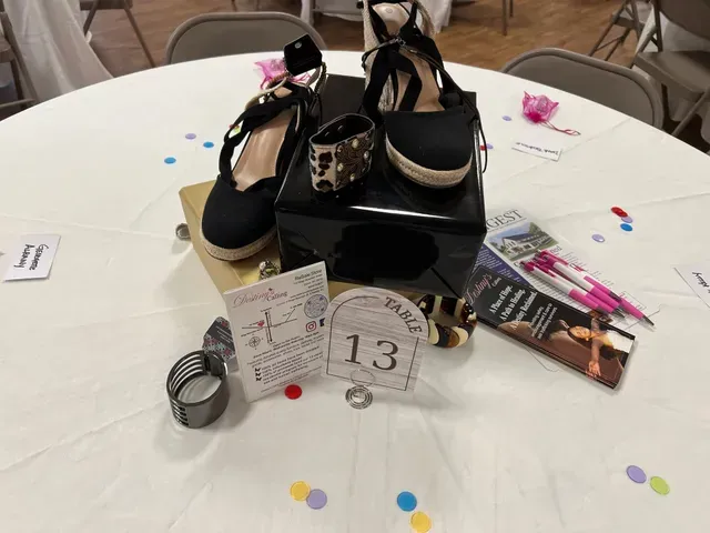 A table centerpiece at an event featuring a pair of black wedge sandals, jewelry, brochures, and a table number 13 card.
