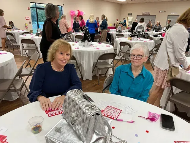 Two people sit at a table in a room decorated with pink, with a silver handbag resting in the foreground.
