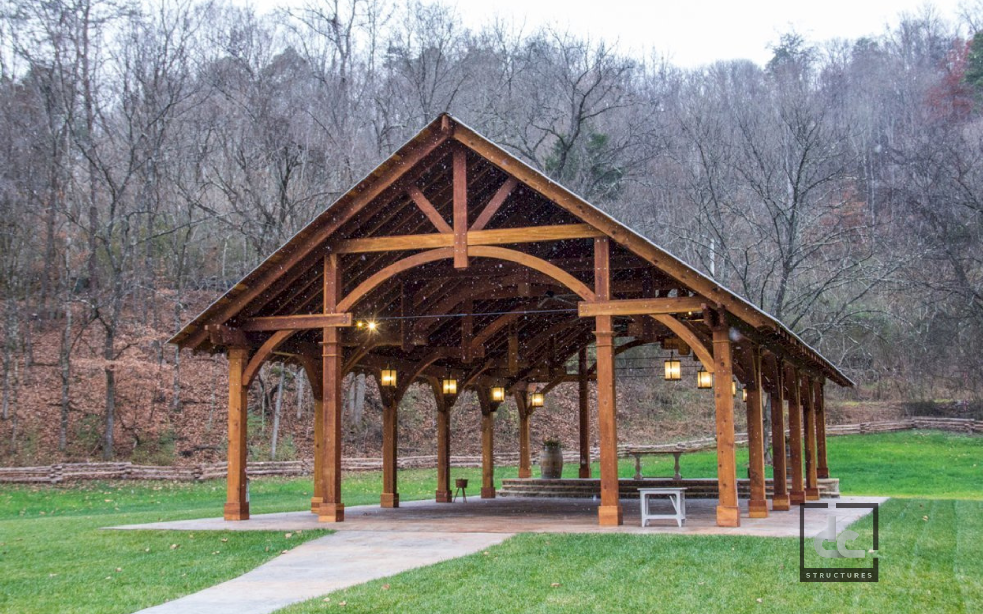 An open-air wooden pavilion with a peaked roof, set on a grassy lawn in front of a wooded hillside.