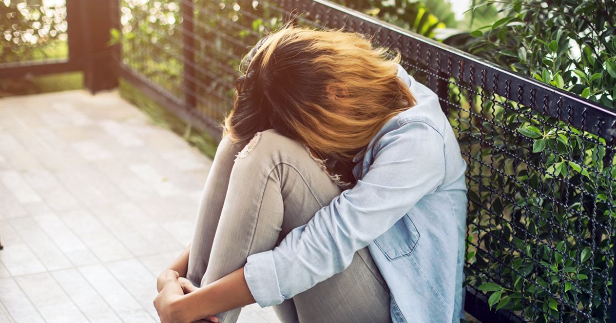 A person in a light blue shirt and jeans sits on a balcony with their head tucked into their knees, appearing distressed.