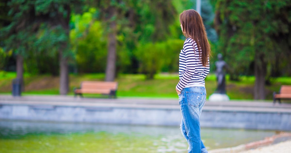 A person with long brown hair, wearing a striped shirt and blue jeans, stands facing away by a pond in a park.