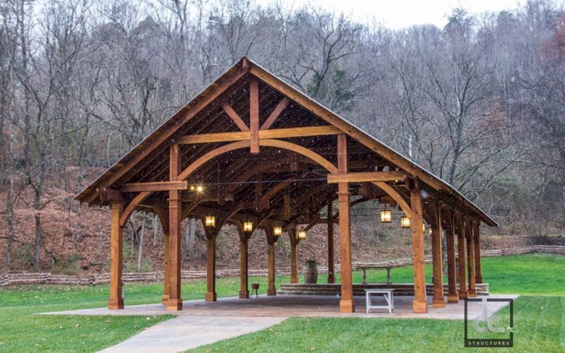 A large, open-air timber-frame pavilion with decorative lighting, situated on a grassy lawn in front of a wooded hillside.