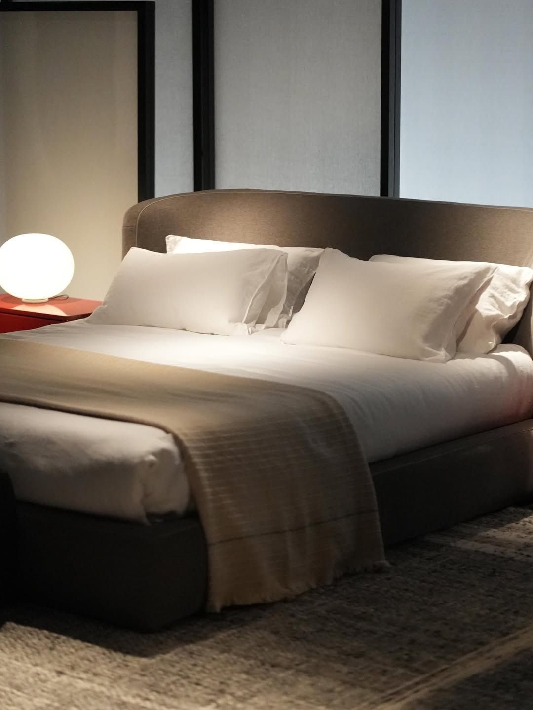 Camera da letto moderna con letto ben rifatto, cuscini bianchi, plaid beige e una lampada da comodino luminosa.