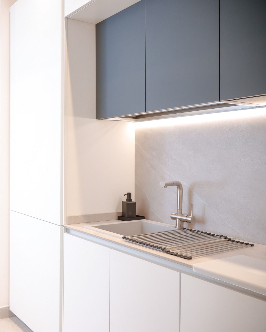 Cucina moderna e minimalista con mobili bianchi, pensili scuri, lavello e illuminazione sottopensile.