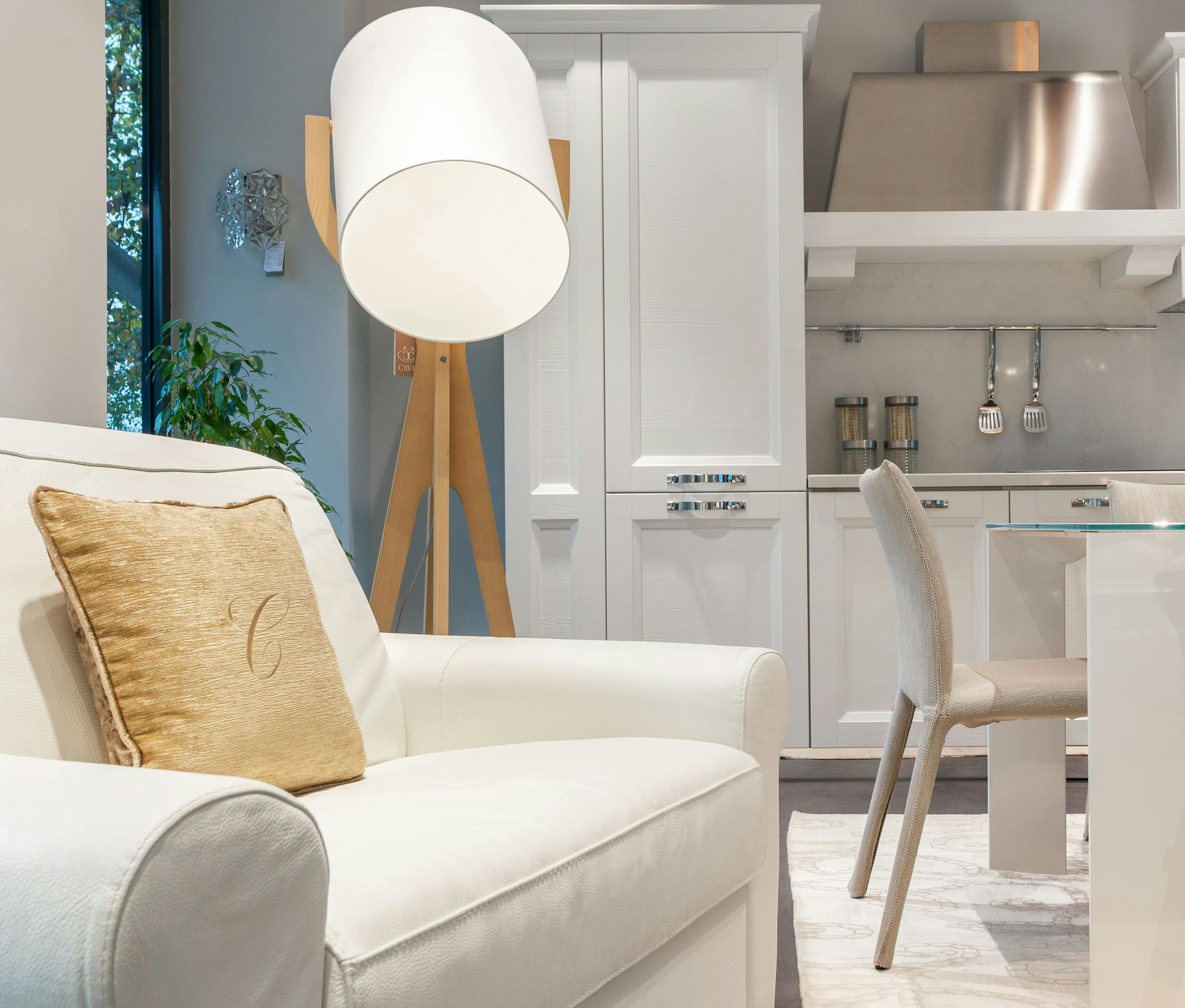 Soggiorno moderno bianco con divano, cuscino dorato, lampada a treppiede e zona pranzo sullo sfondo.