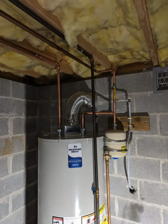 A water heater is sitting in a basement next to a brick wall.