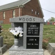 A gray granite tombstone for James Granville and Judy W. Woods with a white flower vase in a cemetery near a brick church.
