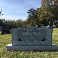 A gray granite tombstone for the Chapman family featuring two etched hearts and dates, set in a sunny cemetery.