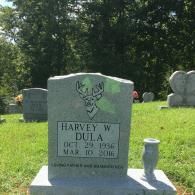 A gray granite tombstone for Harvey W. Dula, featuring an engraved deer, in a sunny cemetery.