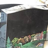 A black headstone featuring a colorful, detailed painting of a log cabin surrounded by trees on a grassy hill.