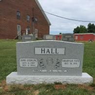 A gray granite headstone for the Hall family in a cemetery, with a brick building and red barn in the background.