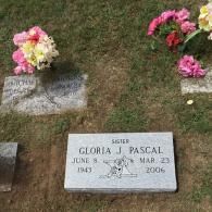 A grave marker for Gloria J. Pascal (1943-2006) in a grassy cemetery, with bouquets of flowers on nearby stones.