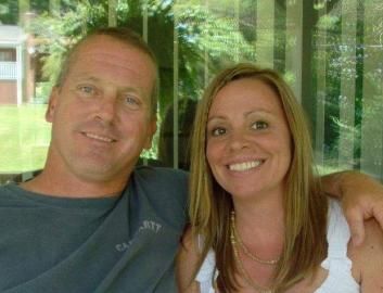 A man in a gray shirt and a woman in a white tank top smile as they pose together in front of a window.