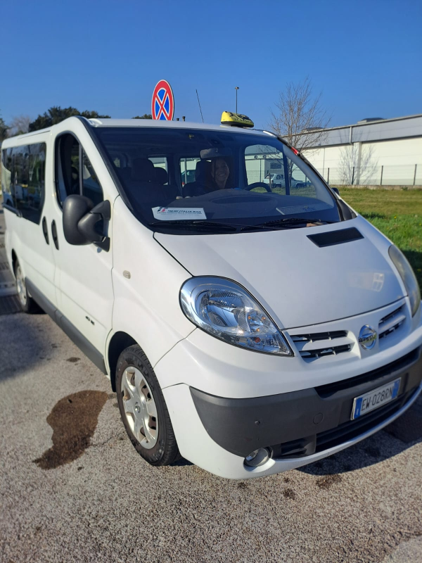 Un furgone taxi Nissan bianco parcheggiato all'aperto in una giornata di sole. L'autista è all'interno.