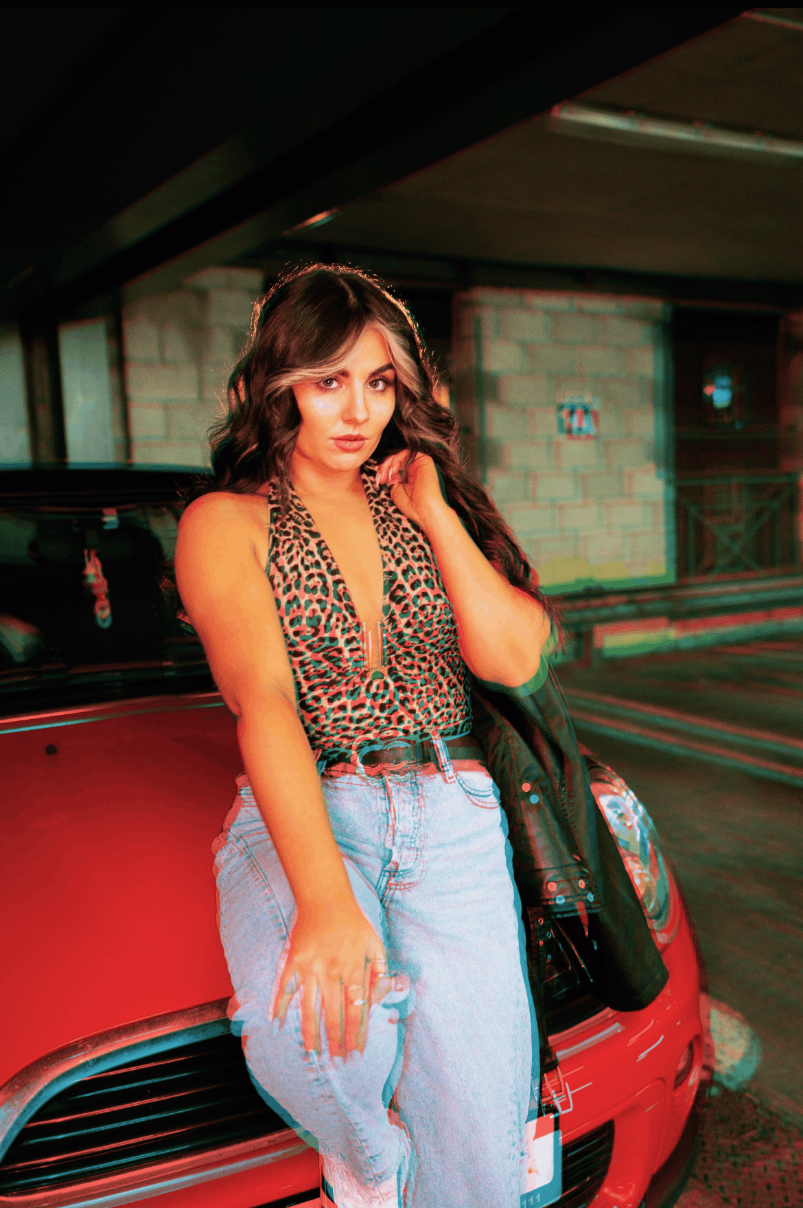 A young girl is standing near a red car in a leopard print top and holding a leather jacket.