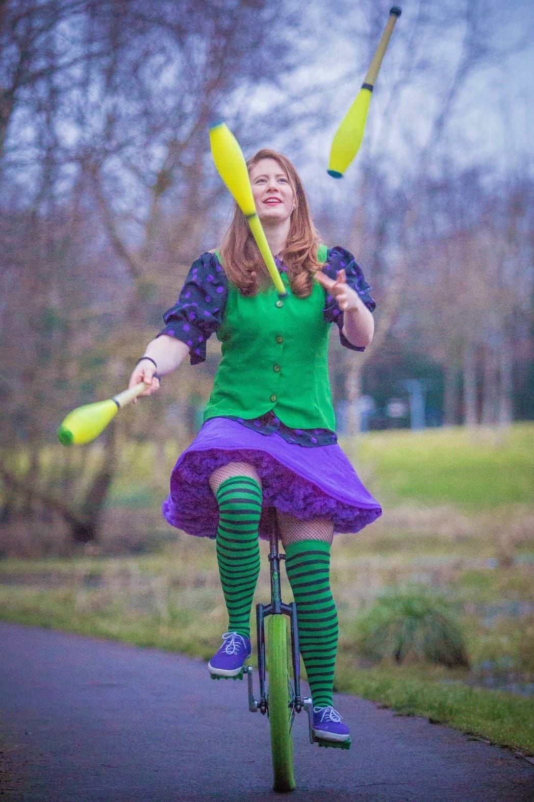 A young woman on a unicycle juggling three colorful juggling clubs.