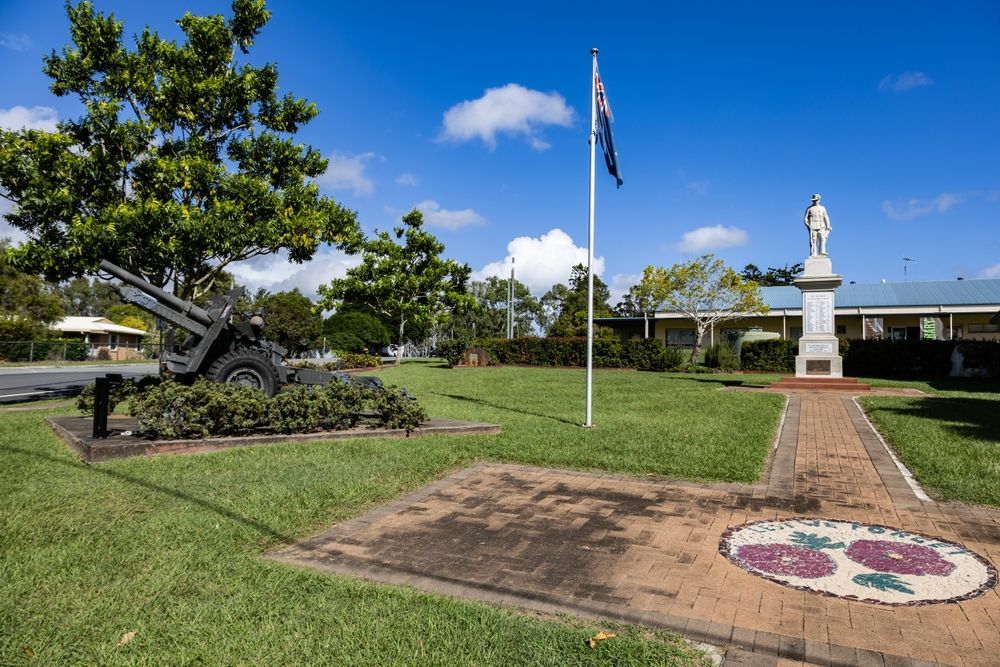 War memorial with cannon, flagpole, statue, and brick path on a grassy lawn under a blue sky.