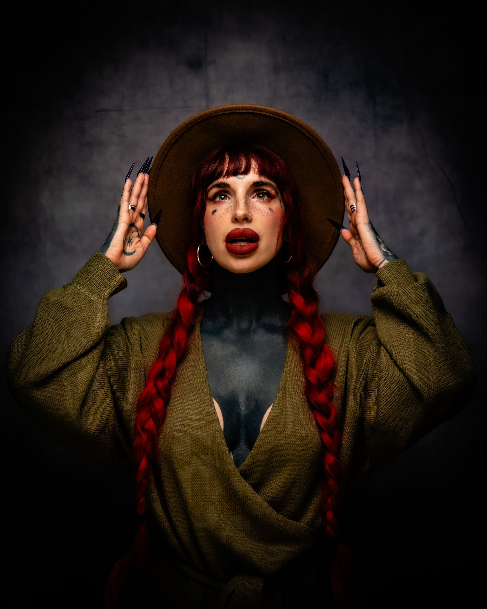Portrait of a person in a wide-brimmed hat and green coat, hands raised beside their face, against a dark background