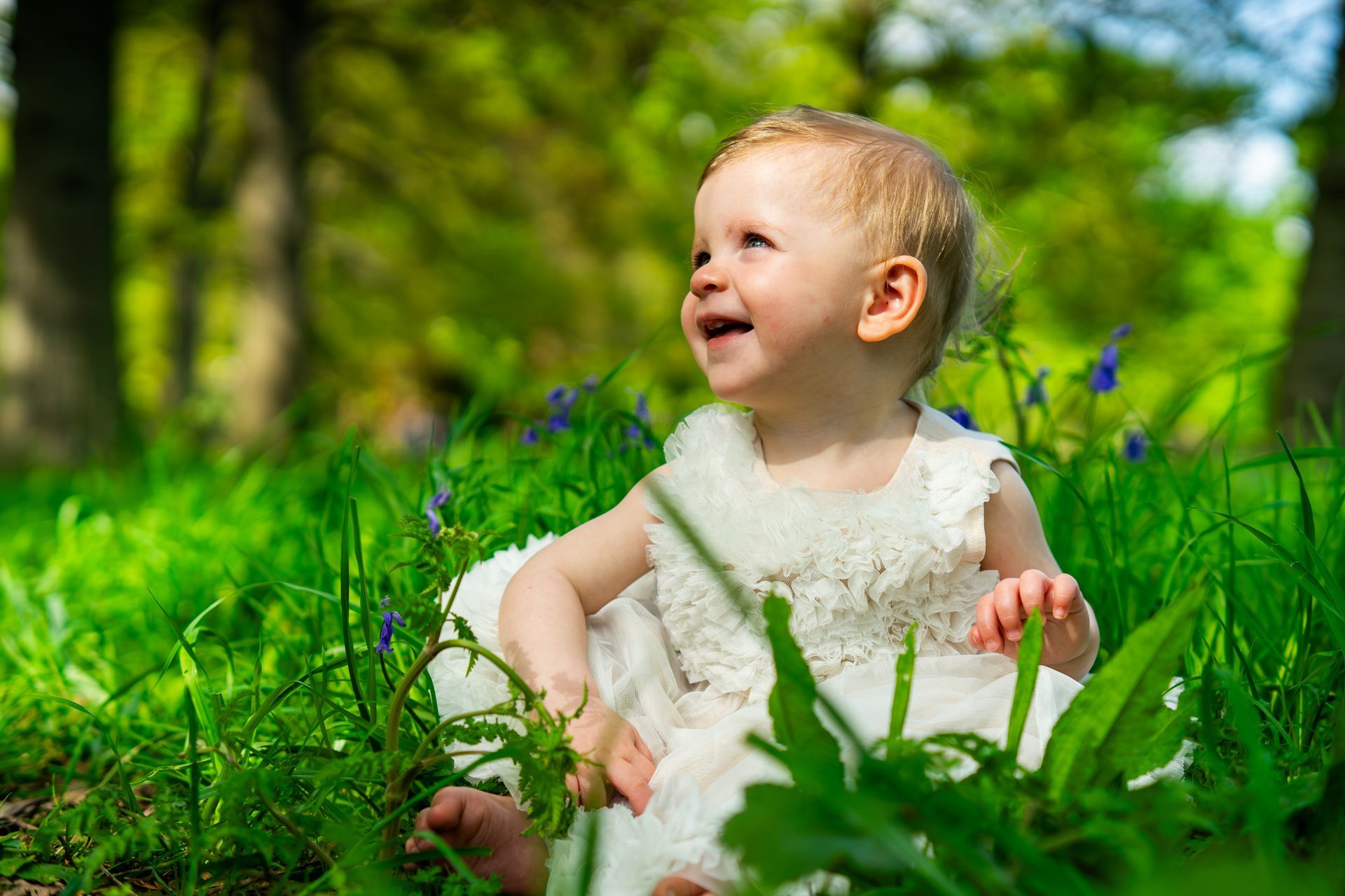 Blond toddler in white dress smiles in grassy field, looking up. Trees in background.