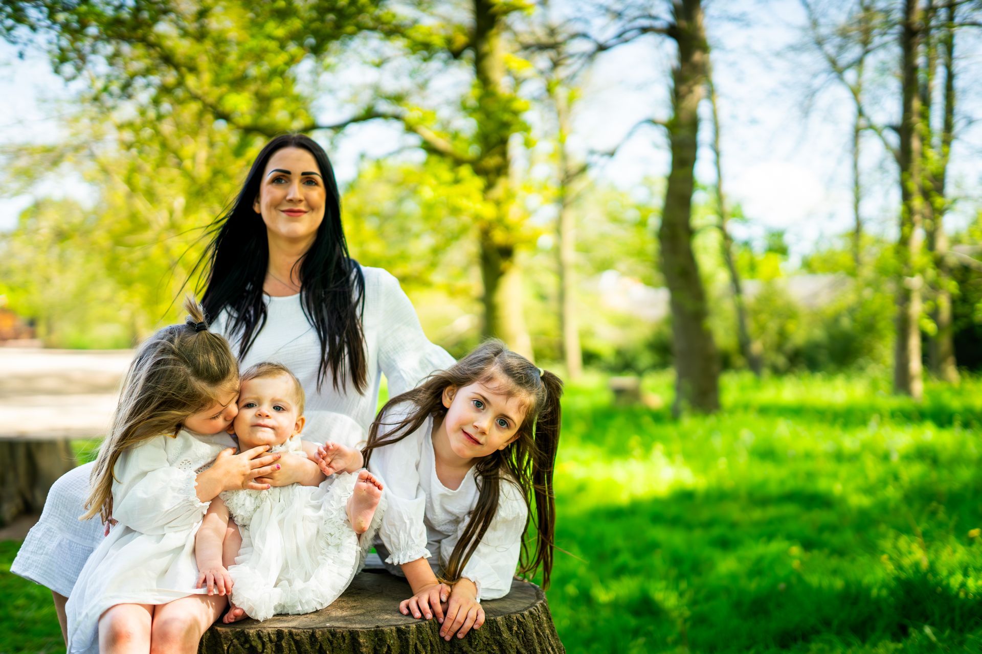 Woman and three young children in white outfits pose outdoors on a tree stump; green grass and trees in background.