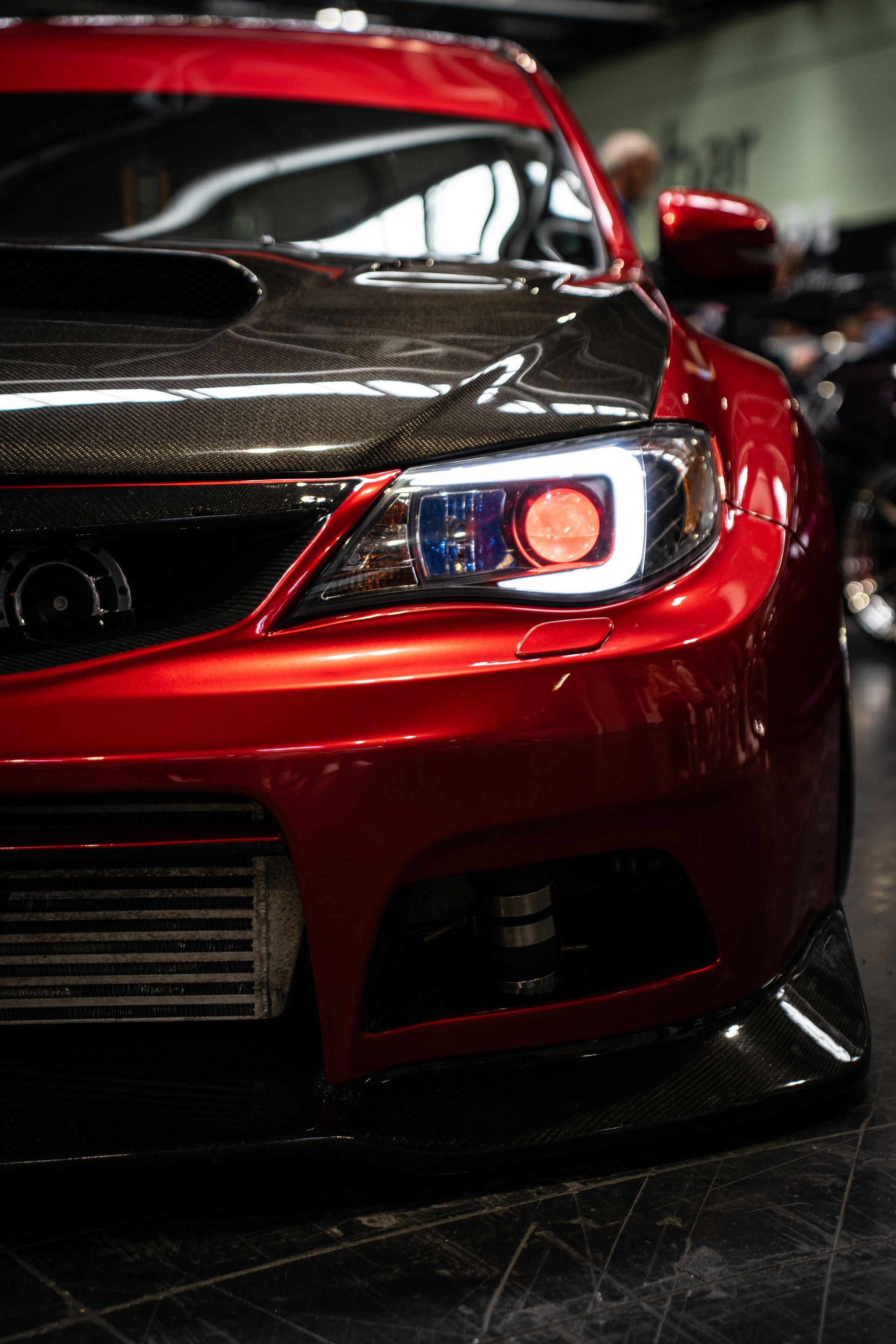 Red Subaru WRX with custom headlights and carbon fiber hood. Indoor setting.