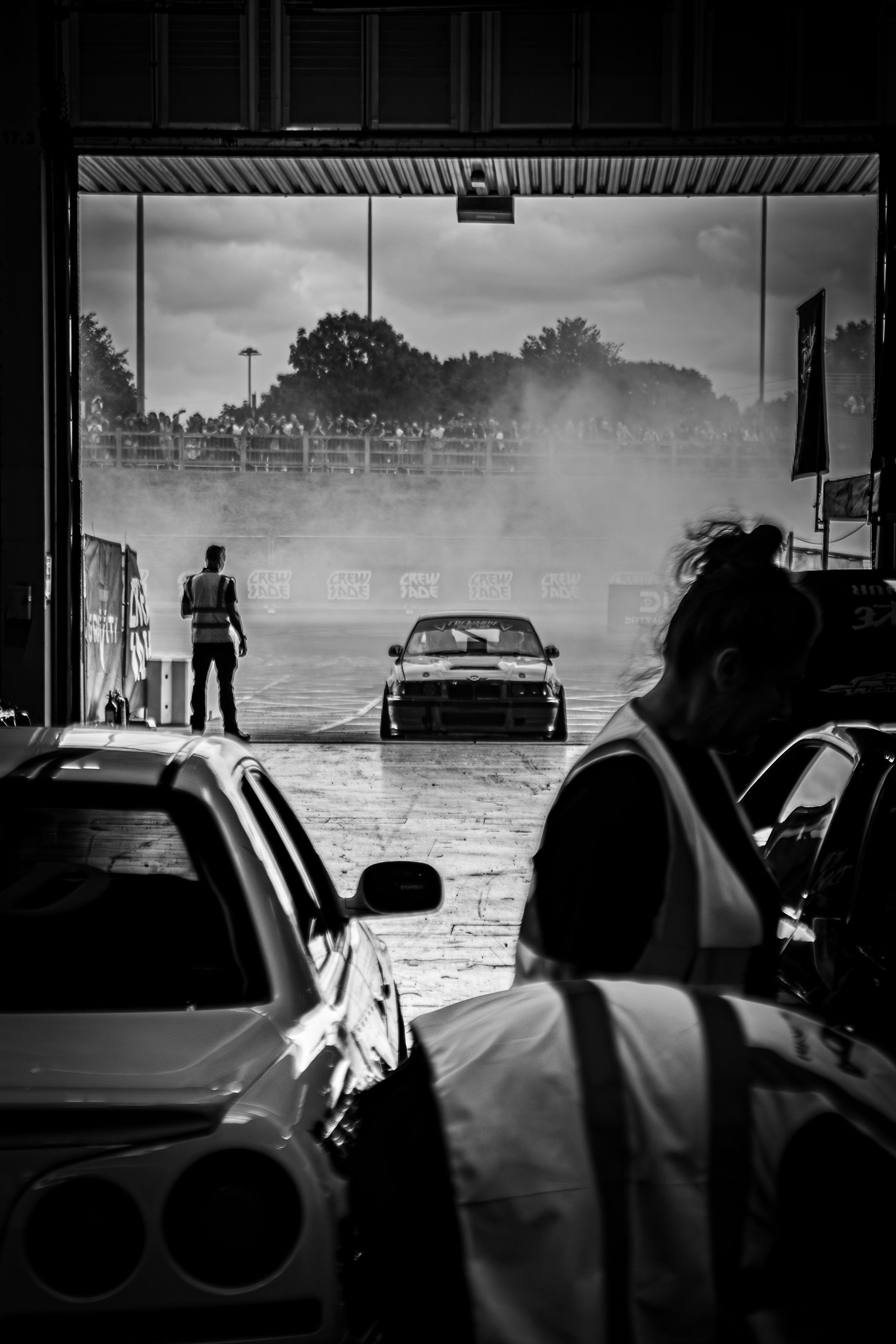 Race car emerging from a garage, generating smoke. Person in a vest watches; another person in the foreground.