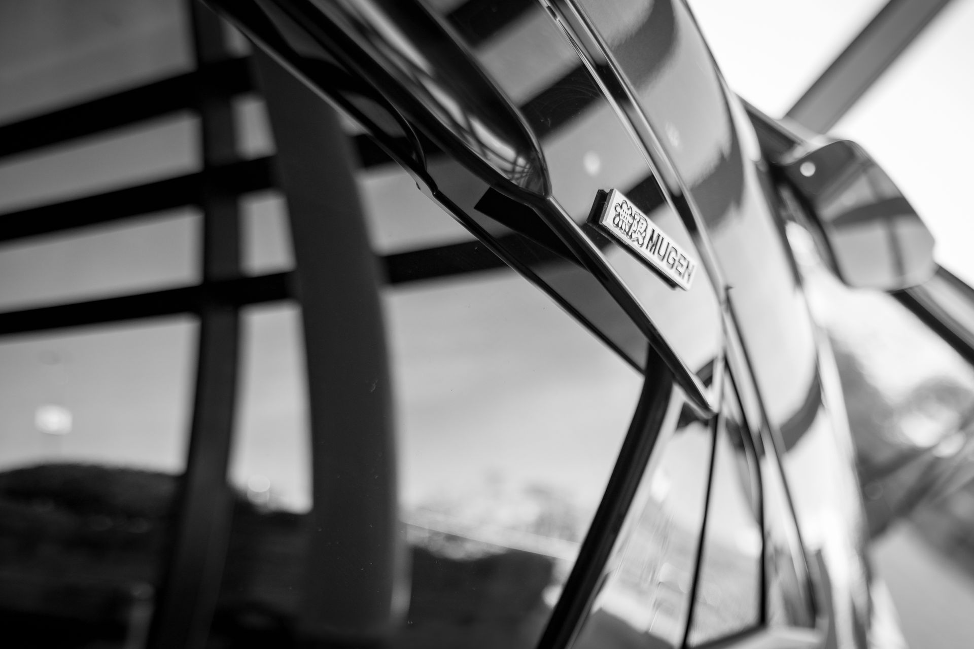 Black and white close-up of a car door with a chrome emblem, reflective glass in the background.