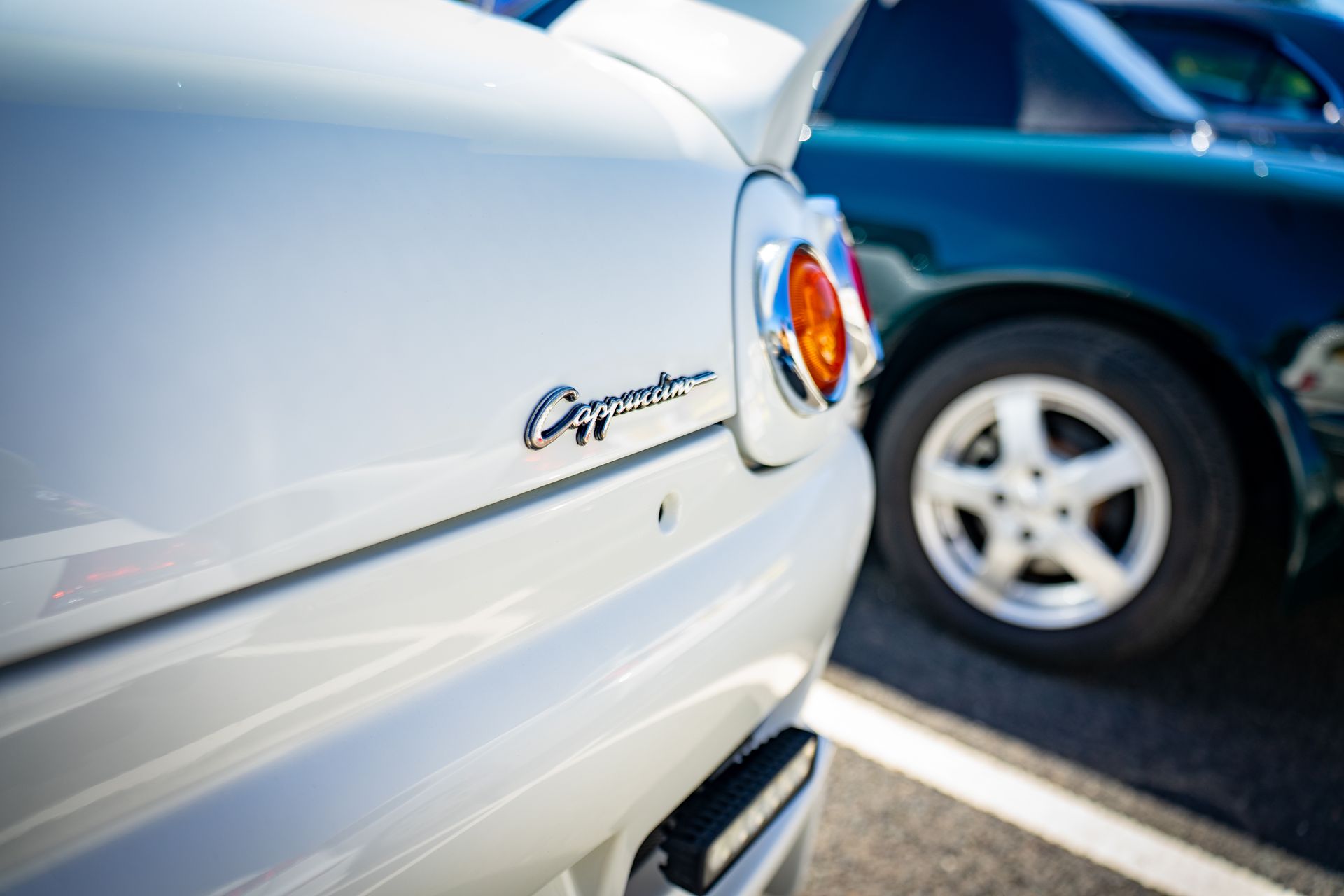 White Cappuccino car rear, chrome lettering, orange taillight, next to a dark green car with a silver wheel.