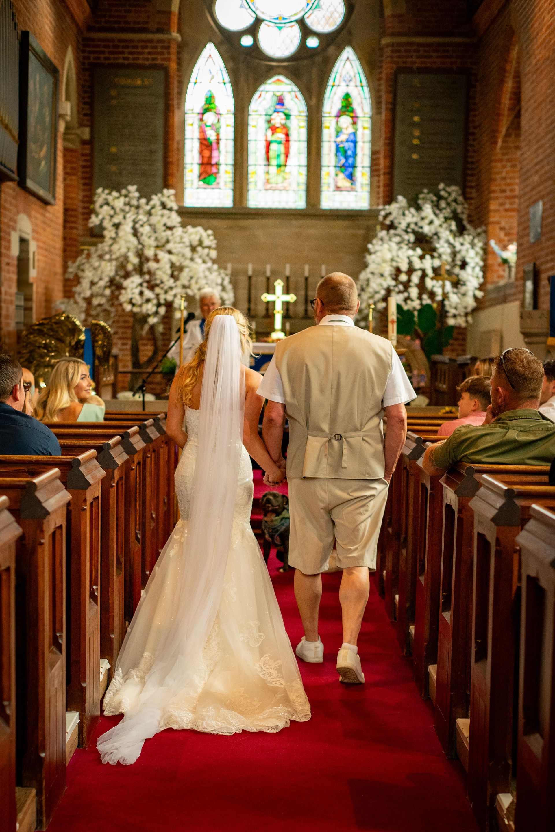 Bride and groom walking down a church aisle after ceremony. Red carpet, stained glass windows, and wedding guests visible.