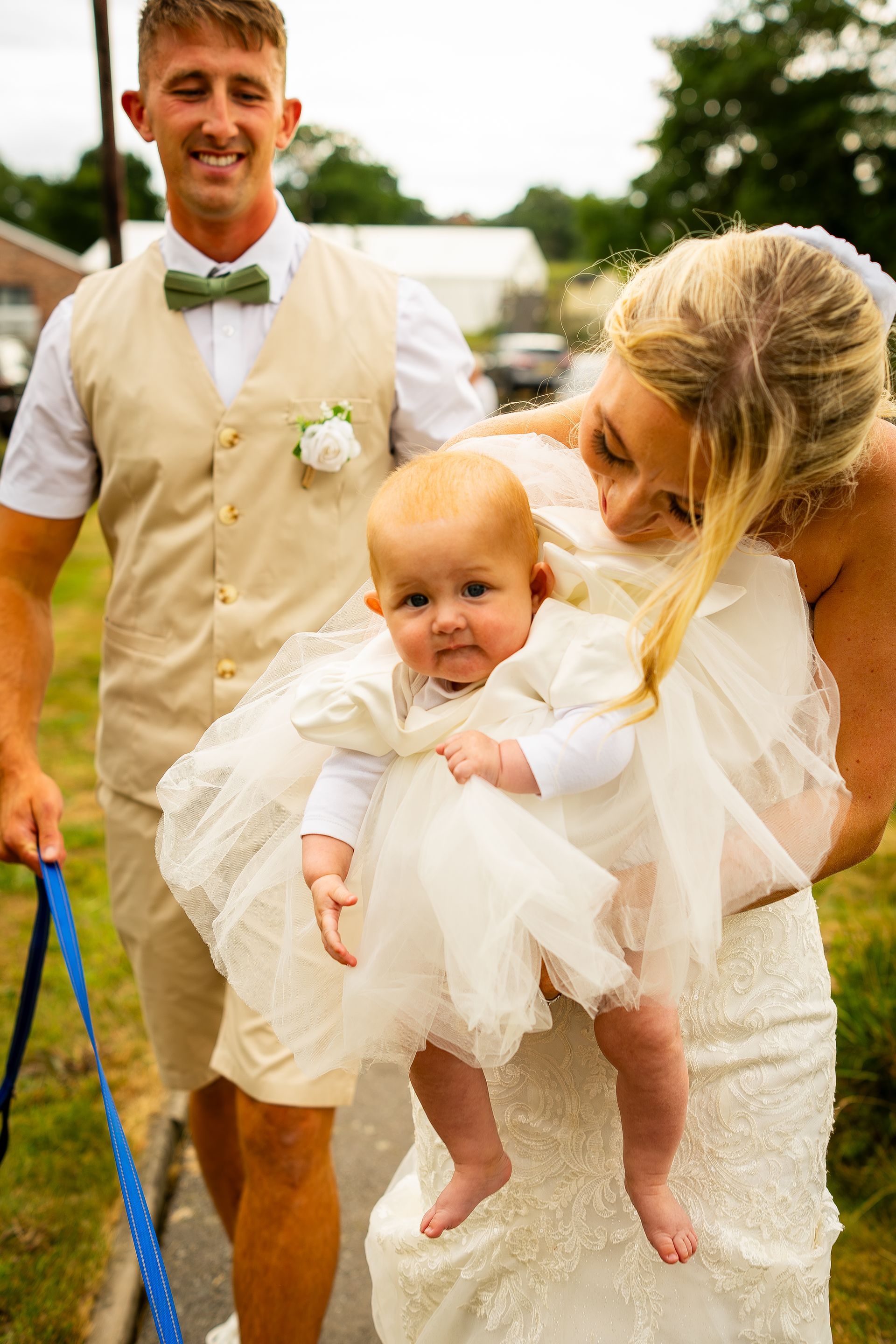 Man in tan suit and woman in wedding dress holding a baby in white dress; outdoors.