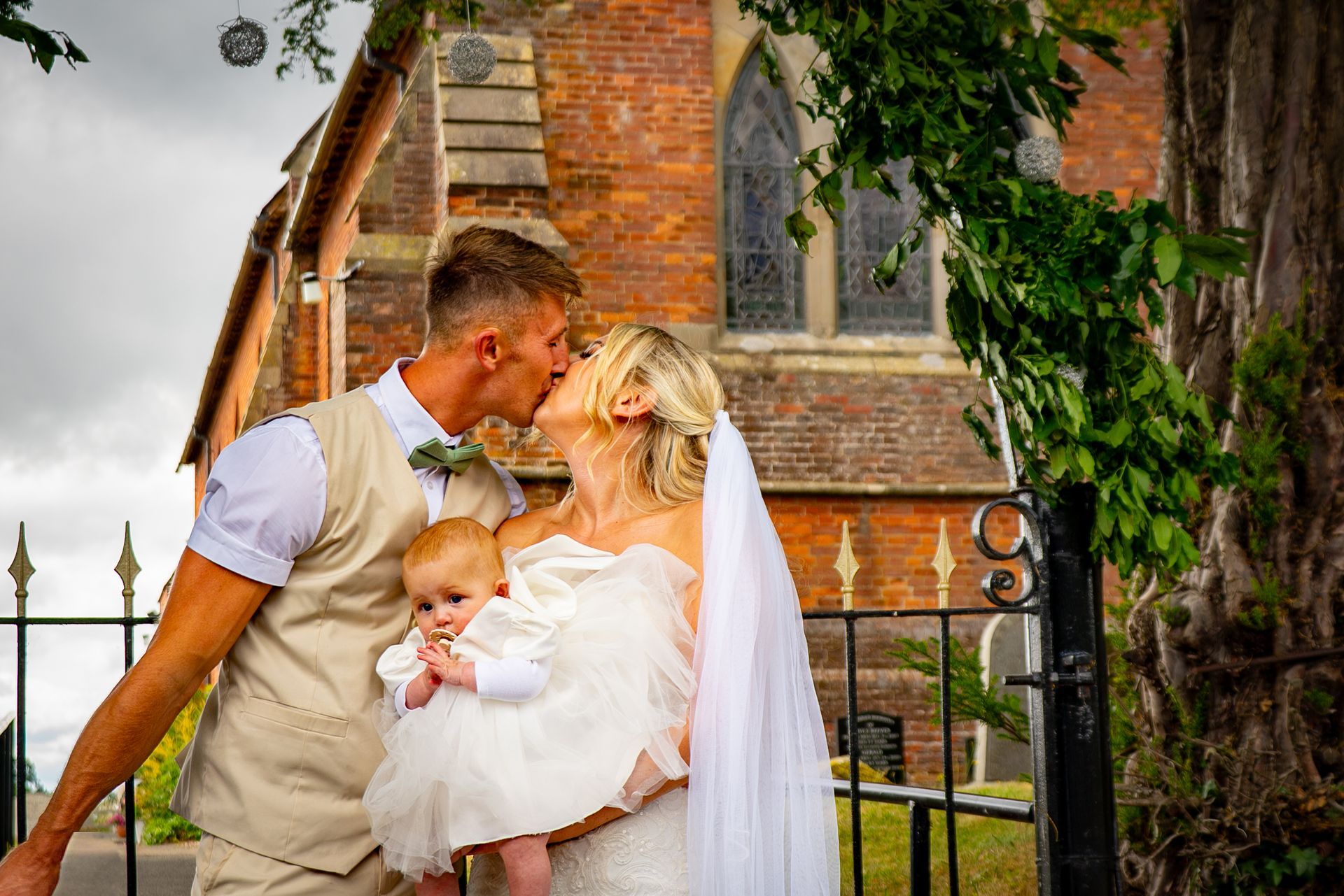 Newlywed couple kissing, holding baby, in front of a brick church.