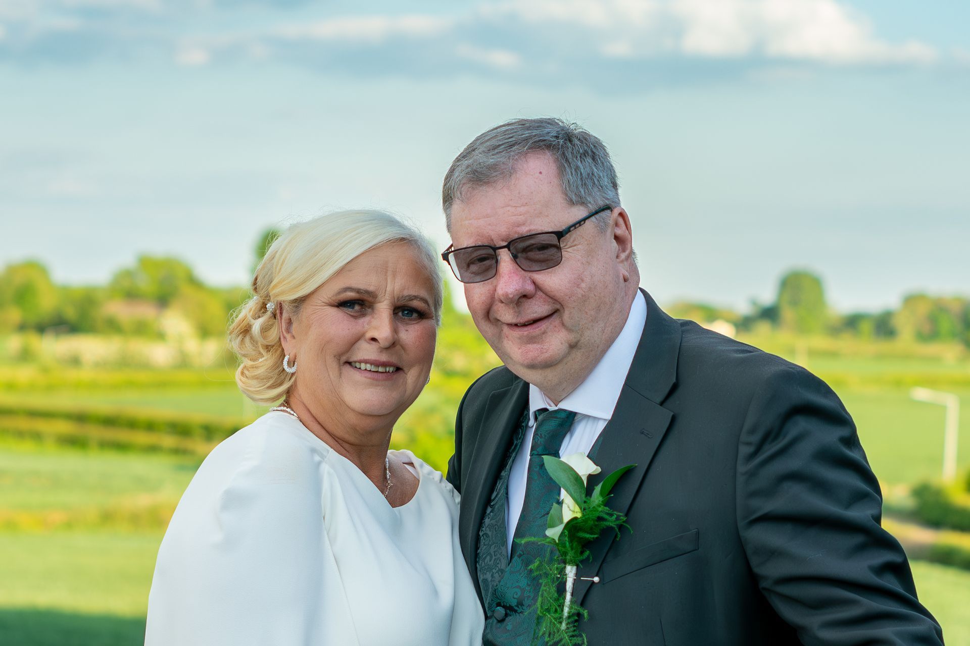 Couple smiles for the camera, outdoors. Woman with blonde hair, white dress, man in suit, green tie. Scenic background.
