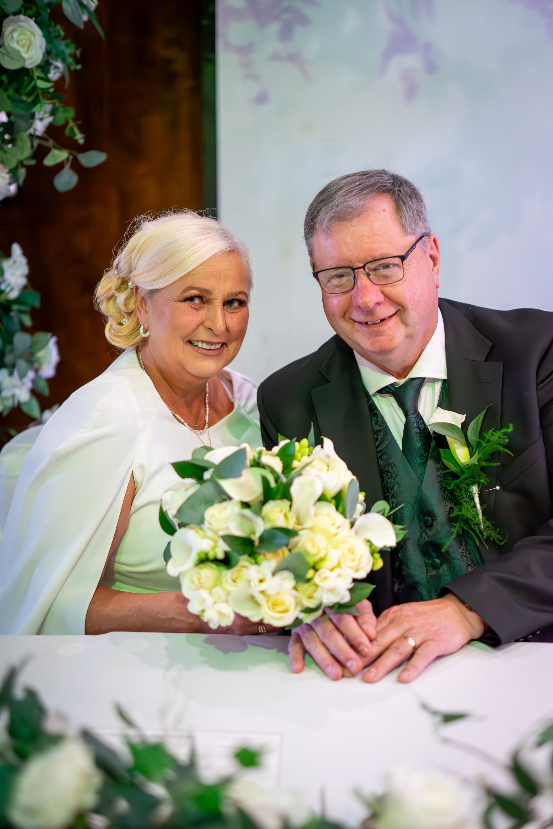 Wedding photo: Smiling couple at a table; woman in white dress holds bouquet; man in suit.