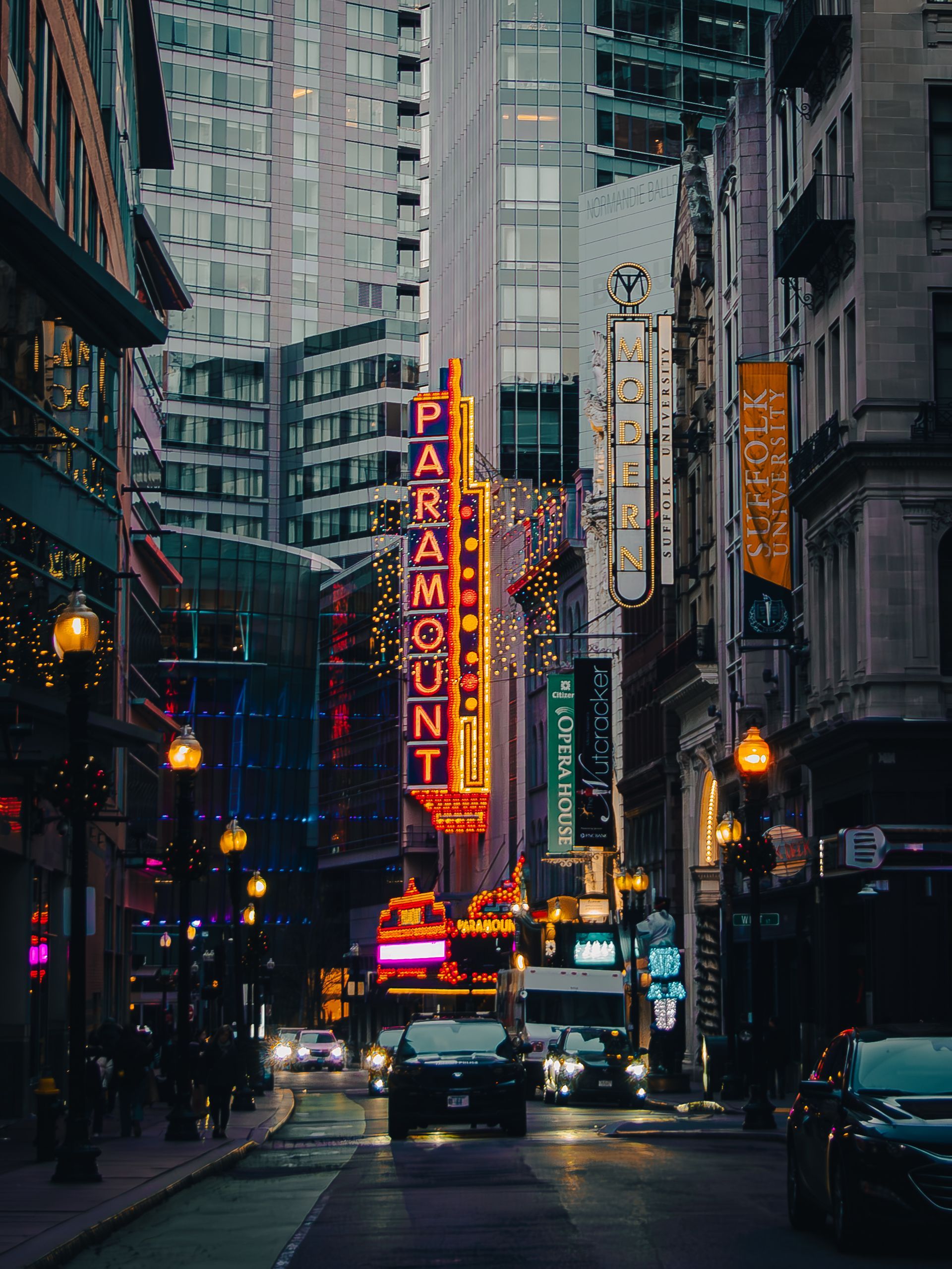 Neon Paramount Theatre sign amidst city buildings at night, cars on the street, vibrant lights.