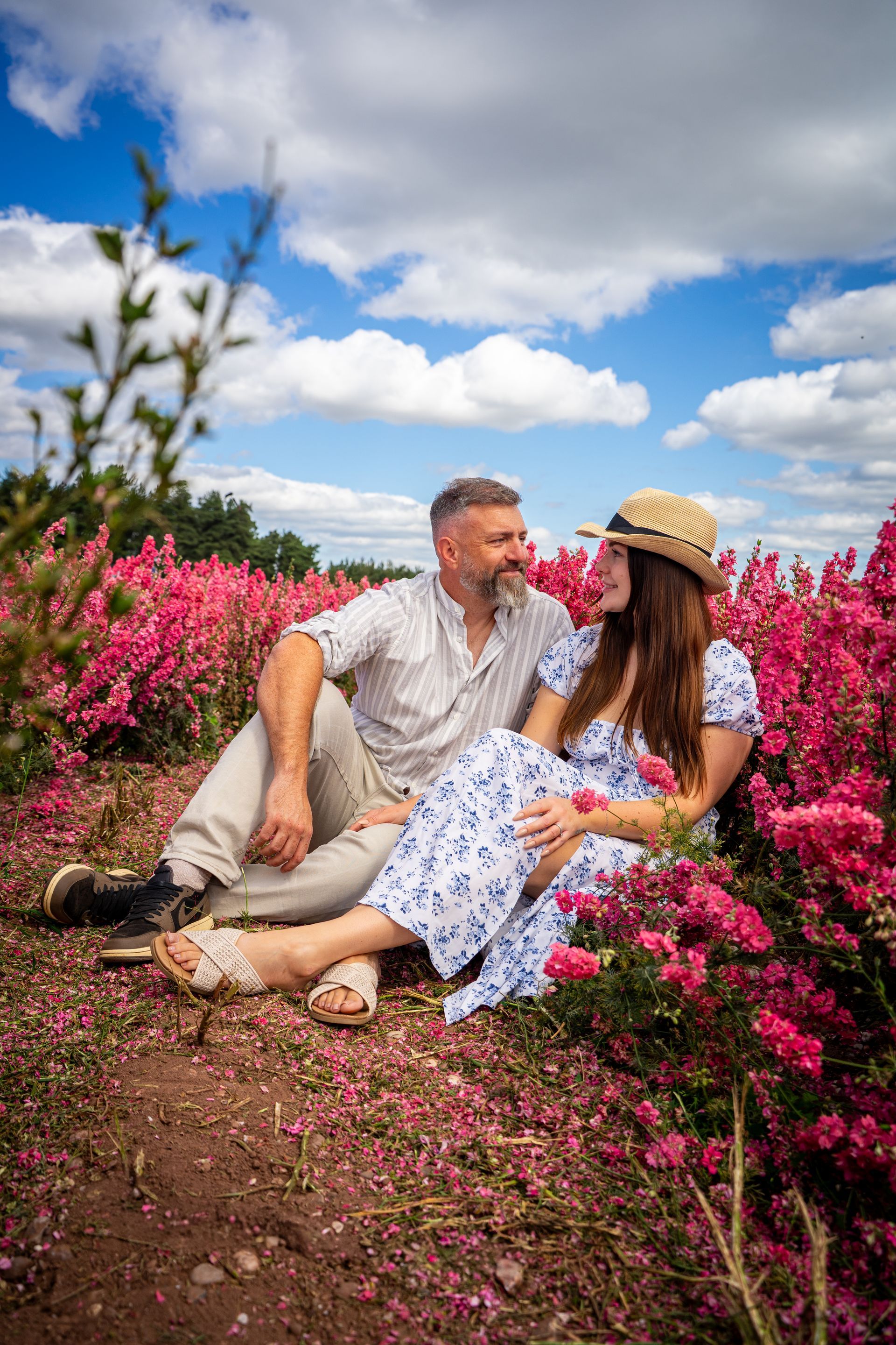 Couple in a field of pink flowers, looking at each other, under a blue sky with clouds.