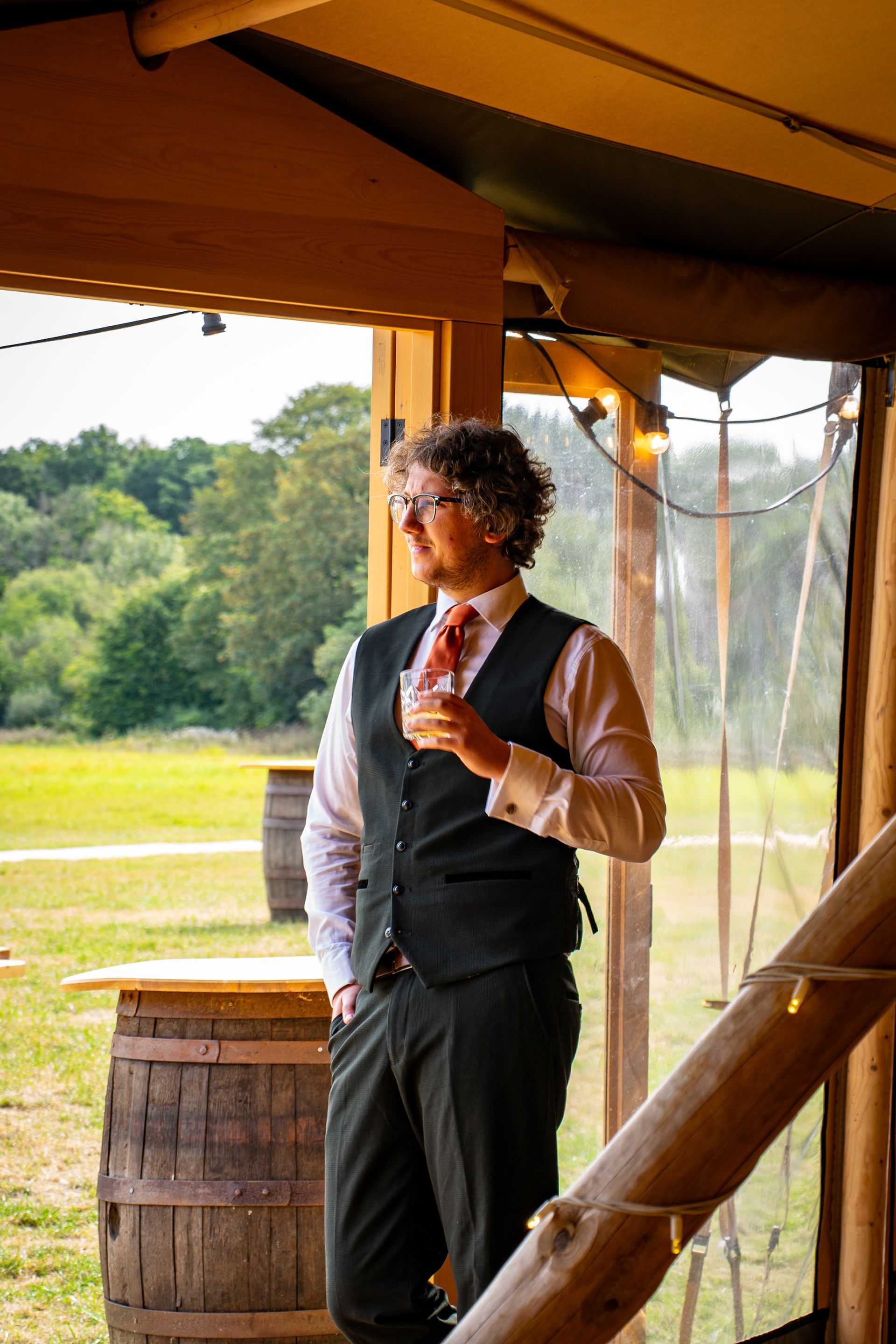 Man in vest and tie holding a drink, looking out over a field from inside a tent.