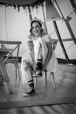 Bride in wedding dress and Converse sneakers laughs, tattoos visible, in a decorated tent.