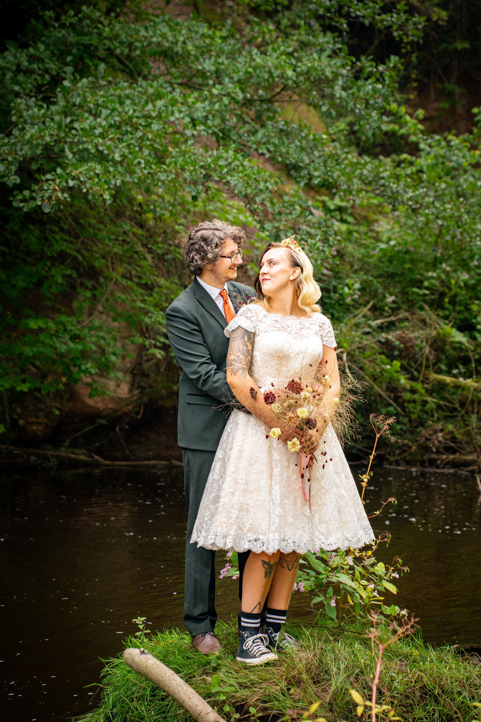 Couple in wedding attire on a small island; green suit, white dress, holding bouquet, smiling, greenery and water background.