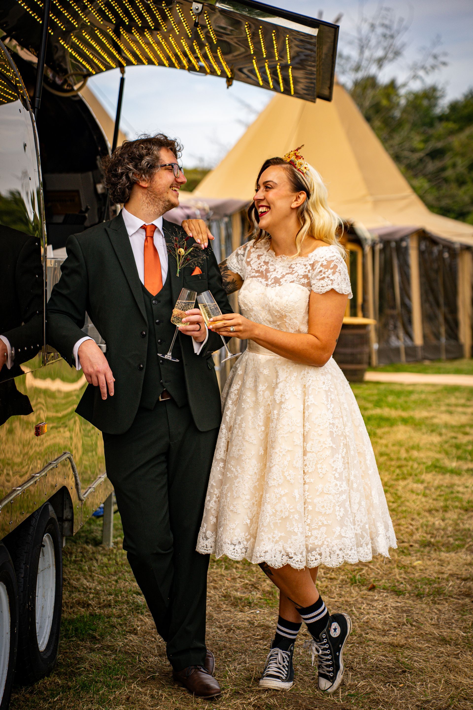 Wedding couple laughing; man in green suit, orange tie; woman in lace dress, skull socks, outdoor setting.