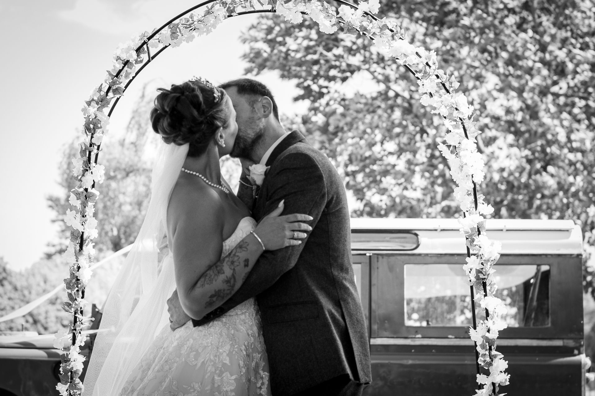 Bride and groom kissing under an arch, side-by-side. Black and white, outdoor setting.