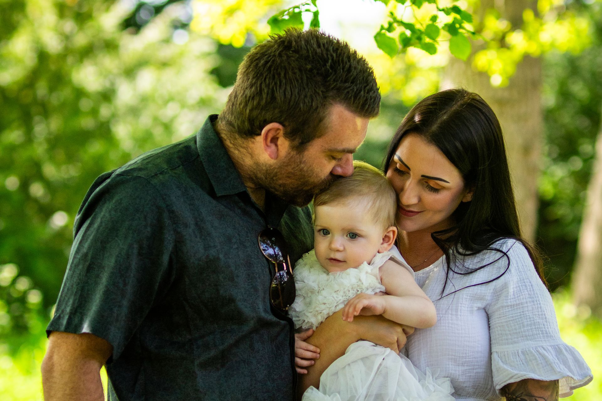 Man kisses baby's head while woman nuzzles the baby. Family outdoors in front of green foliage.