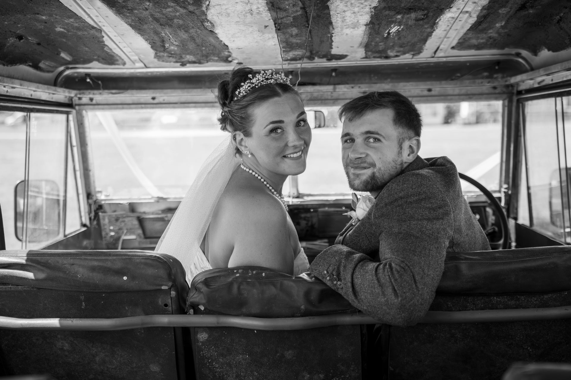 Newlyweds in a vintage car; the bride wears a veil and tiara; the groom smiles; a rustic setting.